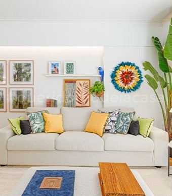Sala com paredes brancas e decoração com cores vivas. Sofá com várias almofadas coloridas por cima, tapete bege e plantas